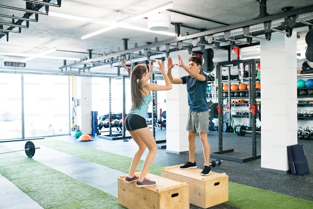 Hermosa pareja joven en forma haciendo ejercicio en el gimnasio moderno, haciendo saltos de caja, chocando los cinco entre sí.