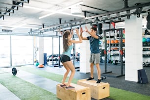 Beautiful young fit couple exercising in modern  gym, doing box jumps, giving high five each other.