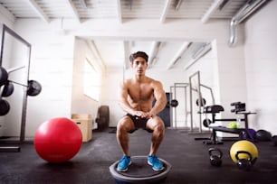 Young fit hispanic man exercising, doing squats on fitness ball in gym gym.