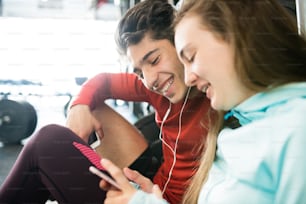 Beautiful young fit couple in modern gym resting, holding smart phone, listening music or watching something.