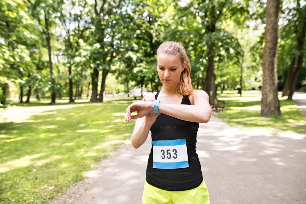 Young woman setting up watch before going for a run in green sunny summer park.
