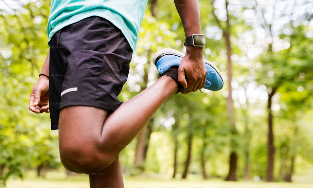 Homme afro-américain méconnaissable dans un parc en train de s’échauffer et d’étirer les bras et les jambes avant l’entraînement.