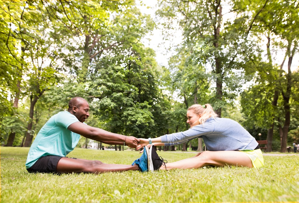 Running couple in park sitting on the grass, warming up and stretching arms and legs before training.