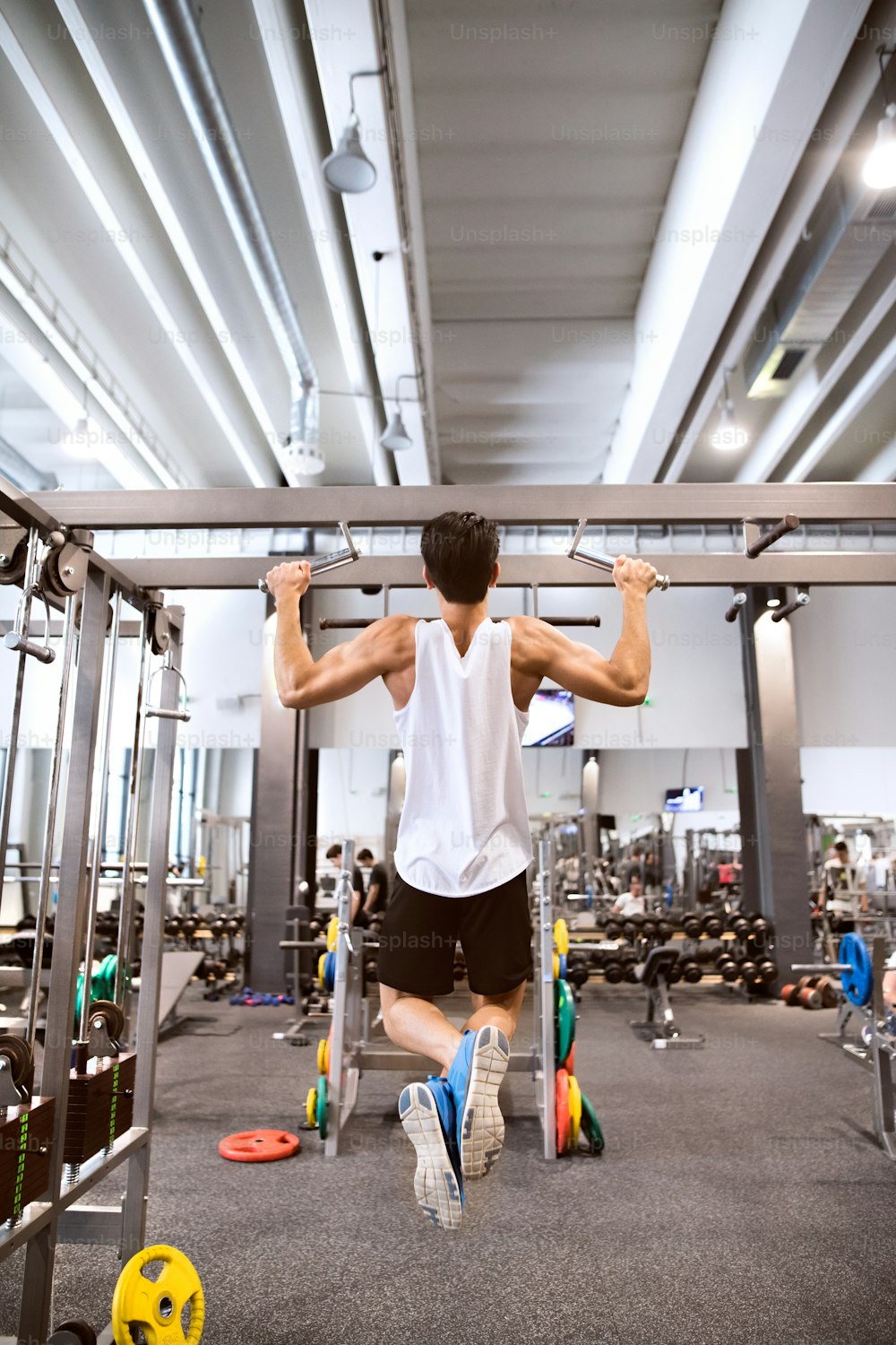 Young hispanic fitness man in gym working out, doing pull-ups on horizontal bar. Rear view.