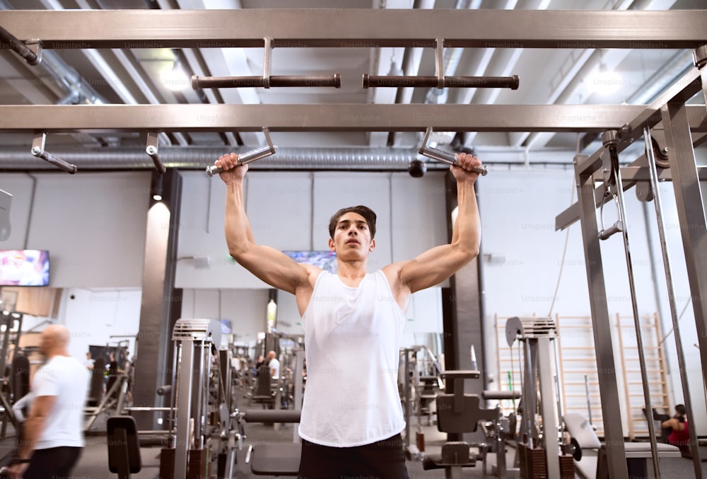 Young hispanic fitness man in gym working out, doing pull-ups on horizontal bar