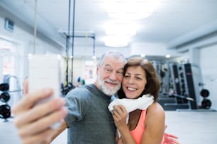 Beautiful fit senior couple in sports clothing in gym resting, taking selfie with smart phone