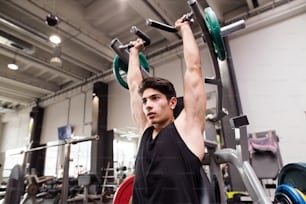 Young fit hispanic man in gym working out on fitness machine, flexing muscles. Bodybuilder training, doing shoulder press.