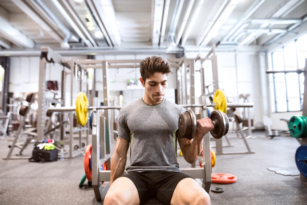 Joven hombre hispano de la aptitud en el gimnasio sentado en el banco, haciendo ejercicio con pesas