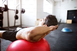 Fit hispanic man at abdominal crunch muscles exercises on red fitness ball during training in fitness gym.