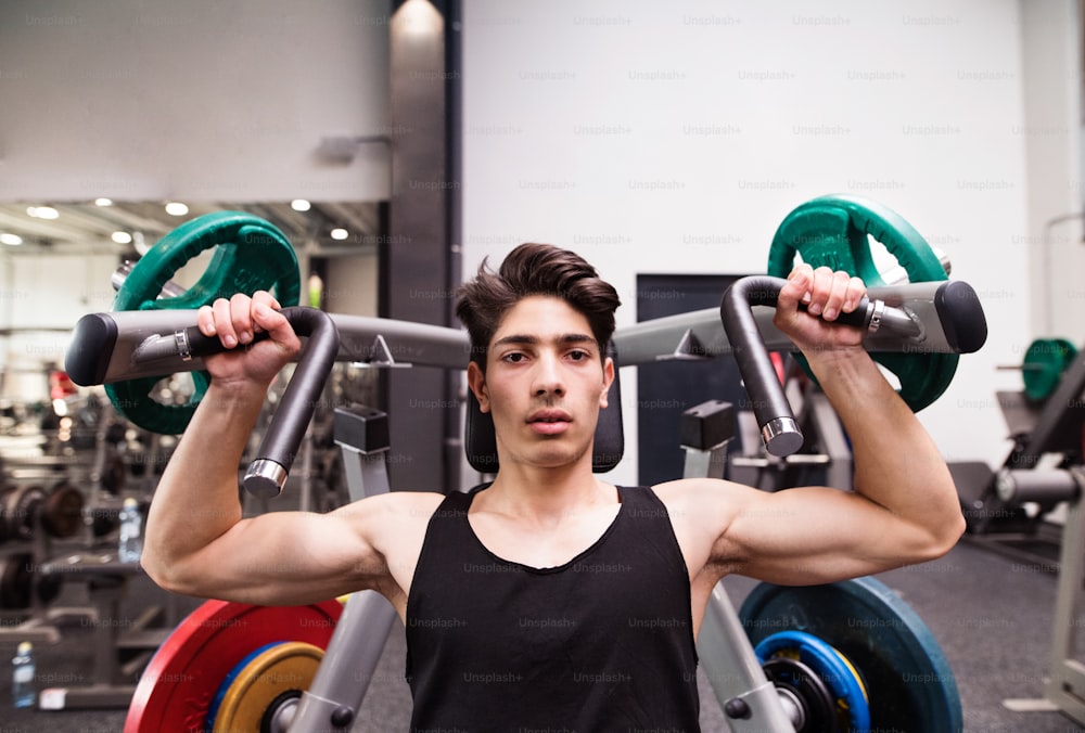 Young fit hispanic man in gym working out on fitness machine, flexing muscles. Bodybuilder training, doing shoulder press.