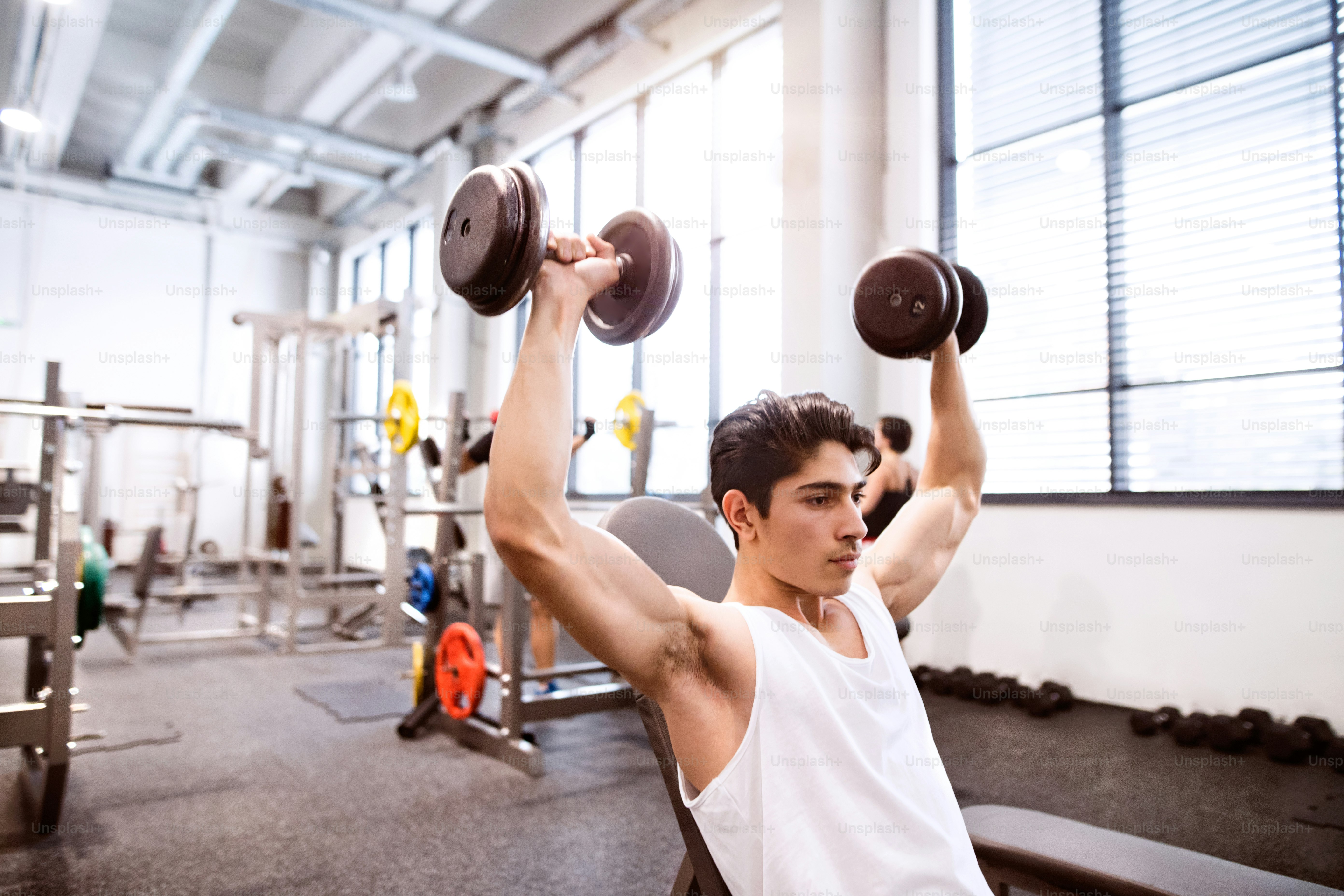Young hispanic fitness man in gym sitting on bench, working out with weights