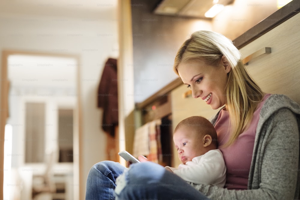 Beautiful young blond mother at home with her little baby son in the arms, sitting on the kitchen floor, holding smart phone, smiling