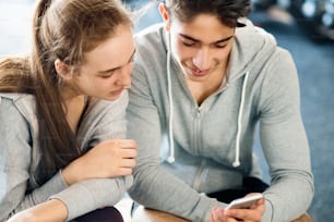 Beautiful young fit couple in modern  gym talking, resting, drinking water, holding smart phone, watching something.