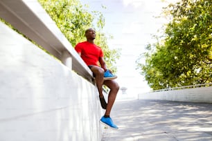 Joven corredor afroamericano en la ciudad sentado en una pared de concreto sosteniendo un zapato, descansando.