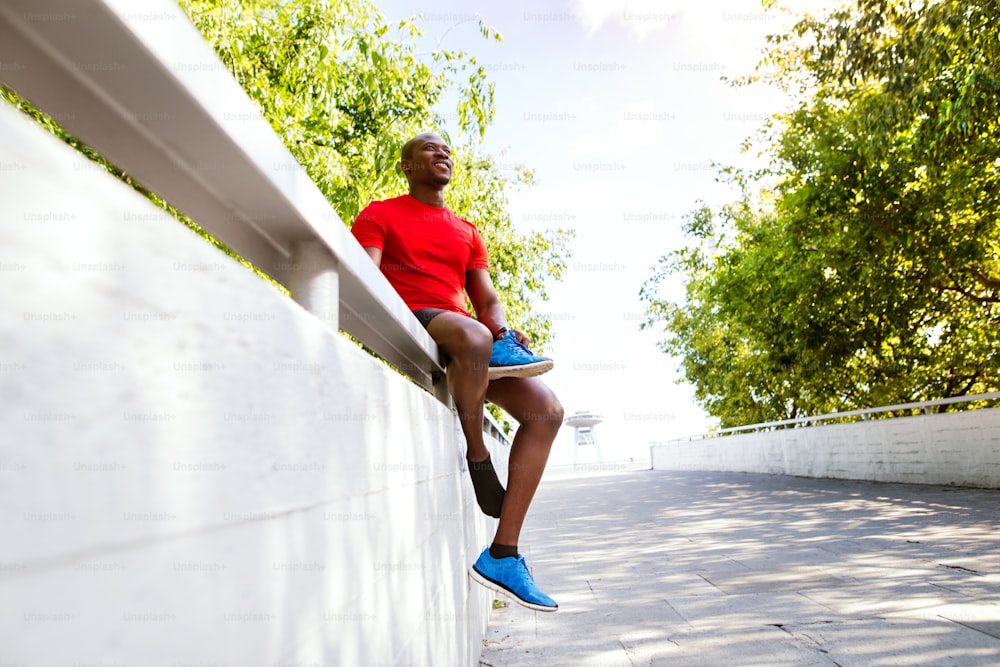 Joven corredor afroamericano en la ciudad sentado en una pared de concreto sosteniendo un zapato, descansando.