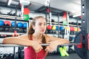 Beautiful young fit woman in gym at the heavy barbell, resting, earphones in her ears, listening music