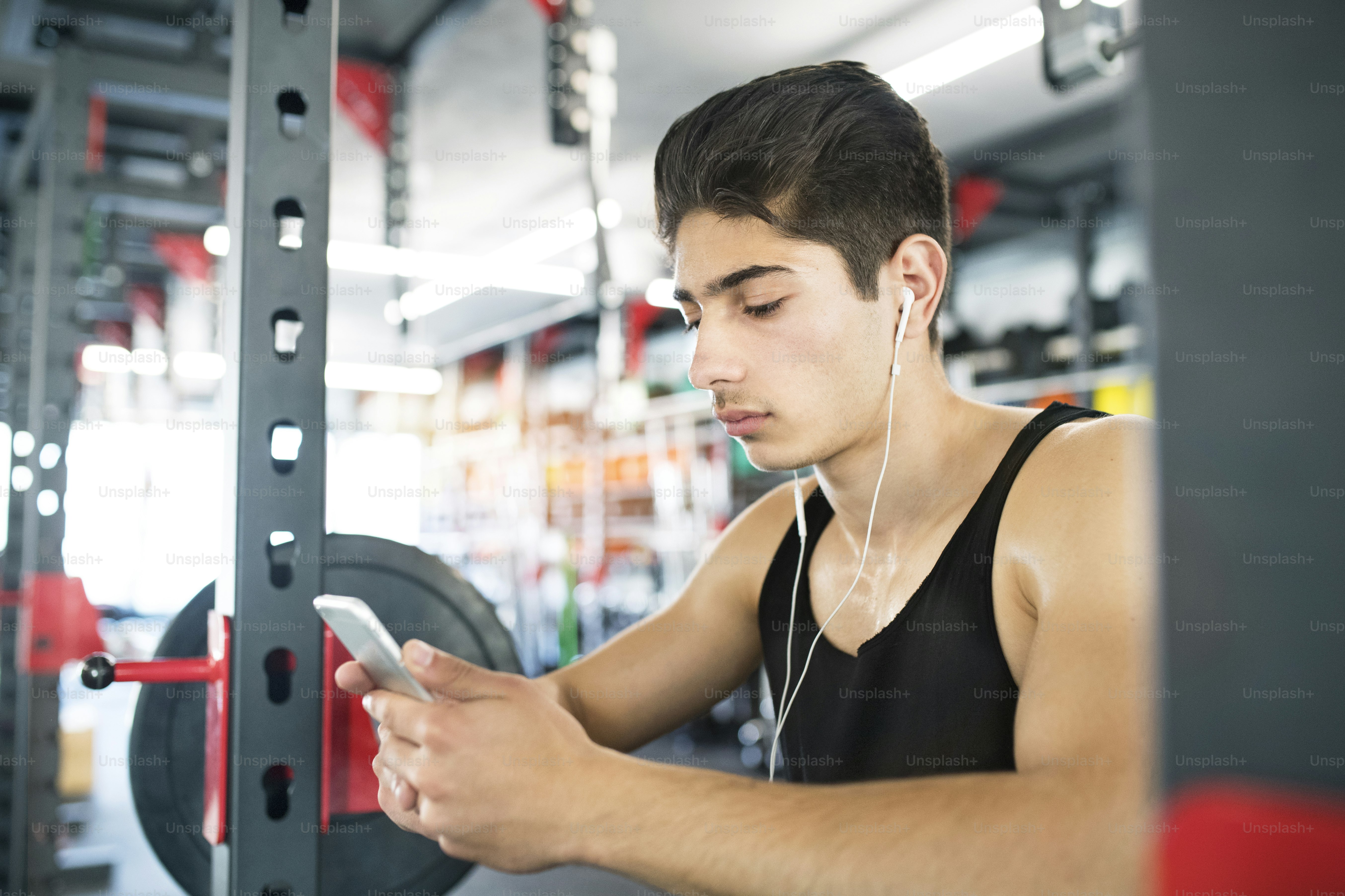 Hombre hispano de la aptitud en el gimnasio descansando, sosteniendo el teléfono inteligente, auriculares en sus oídos escuchando música