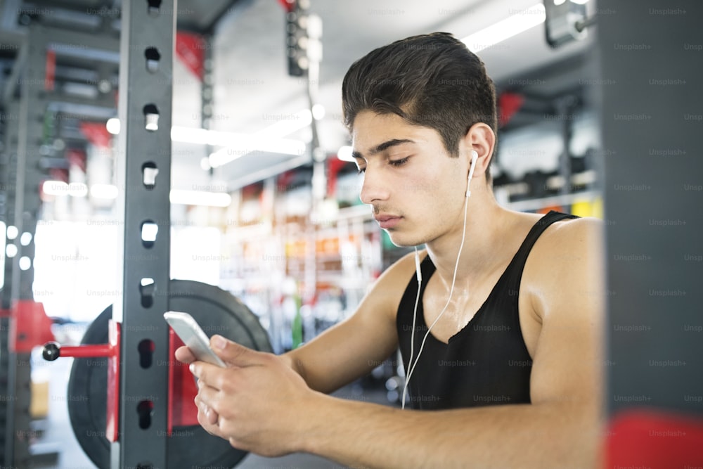 Hispanic fitness man in gym resting, holding smart phone, earphones in his ears listening music