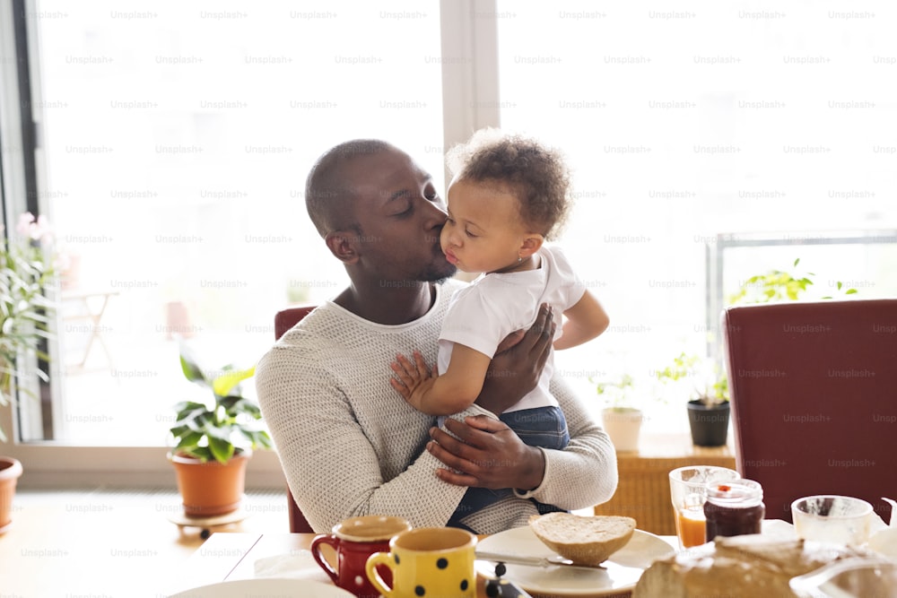 어린 딸과 함께 집에 있는 젊은 아프리카계 미국인 아버지는 그녀를 팔에 안고 뺨에 키스하고 함께 아침을 먹습니다.