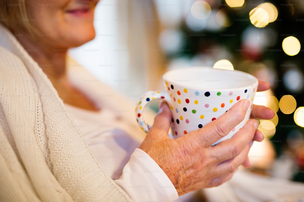 Unrecognizable senior woman sitting on couch in front of illuminated Christmas tree inside in her house enjoying cup of tea or coffee, smiling.