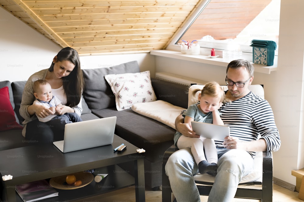 Beautiful young parents at home with children and gadgets. Mother and son with laptop and father with daugter watching something on tablet.