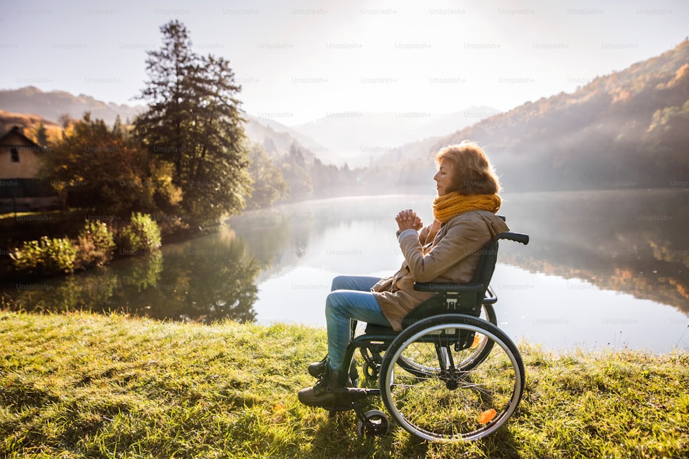 秋の自然の中で車椅子に乗った年配の女性。早朝、湖畔で祈りを捧げる女性。