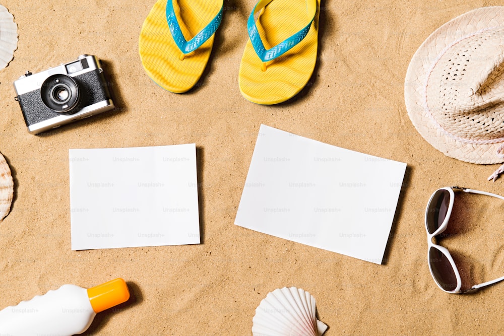 Summer vacation composition with pair of yellow flip flop sandals, hat, sunglasses, sun cream, empty paper sheet and other stuff on a beach. Sand background, studio shot, flat lay. Copy space.
