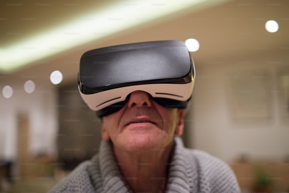 Senior man in gray sweater at home in his living room in the evening wearing virtual reality goggles, reaching out with his arms