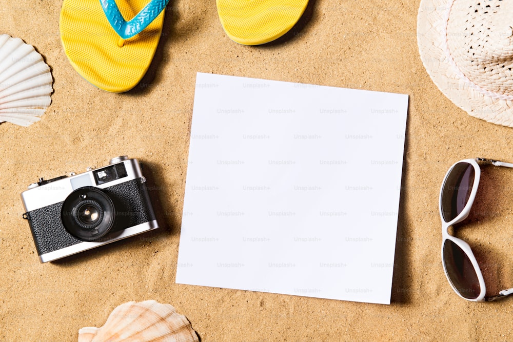 黄色いビーチサンダル、帽子、サングラス、ビンテージカメラ、空の紙シートなどをビーチに置いた夏休みの構図。砂の背景、スタジオショット、フラットレイ。スペースをコピーします。