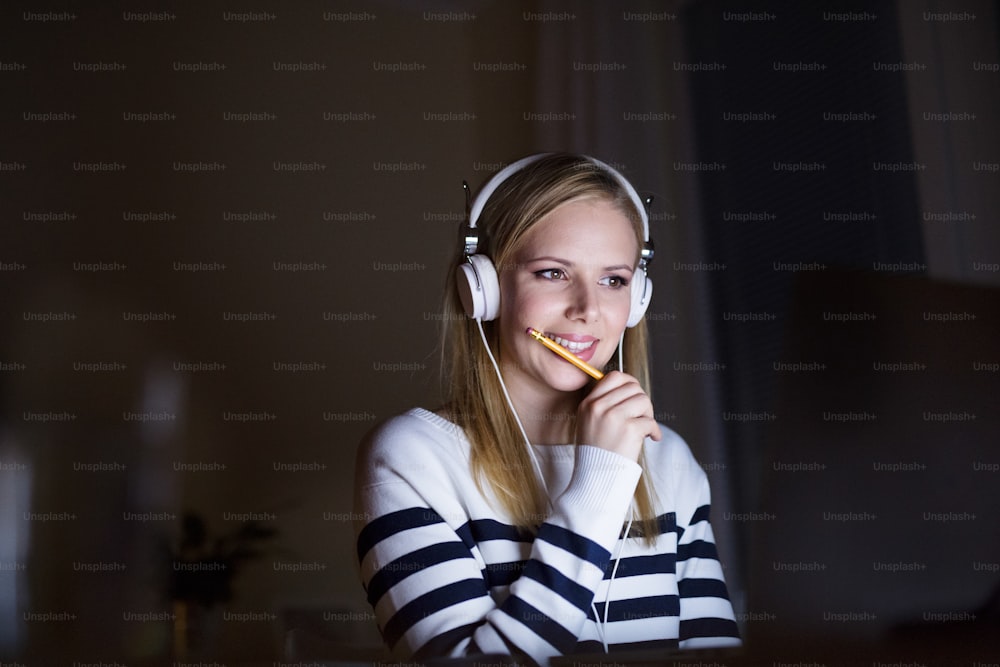 Young beautiful woman sitting at desk at night, holding pencil, headphones on head, smiling.