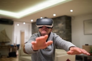 Senior man in gray sweater at home in his living room in the evening wearing virtual reality goggles, reaching out with his arms