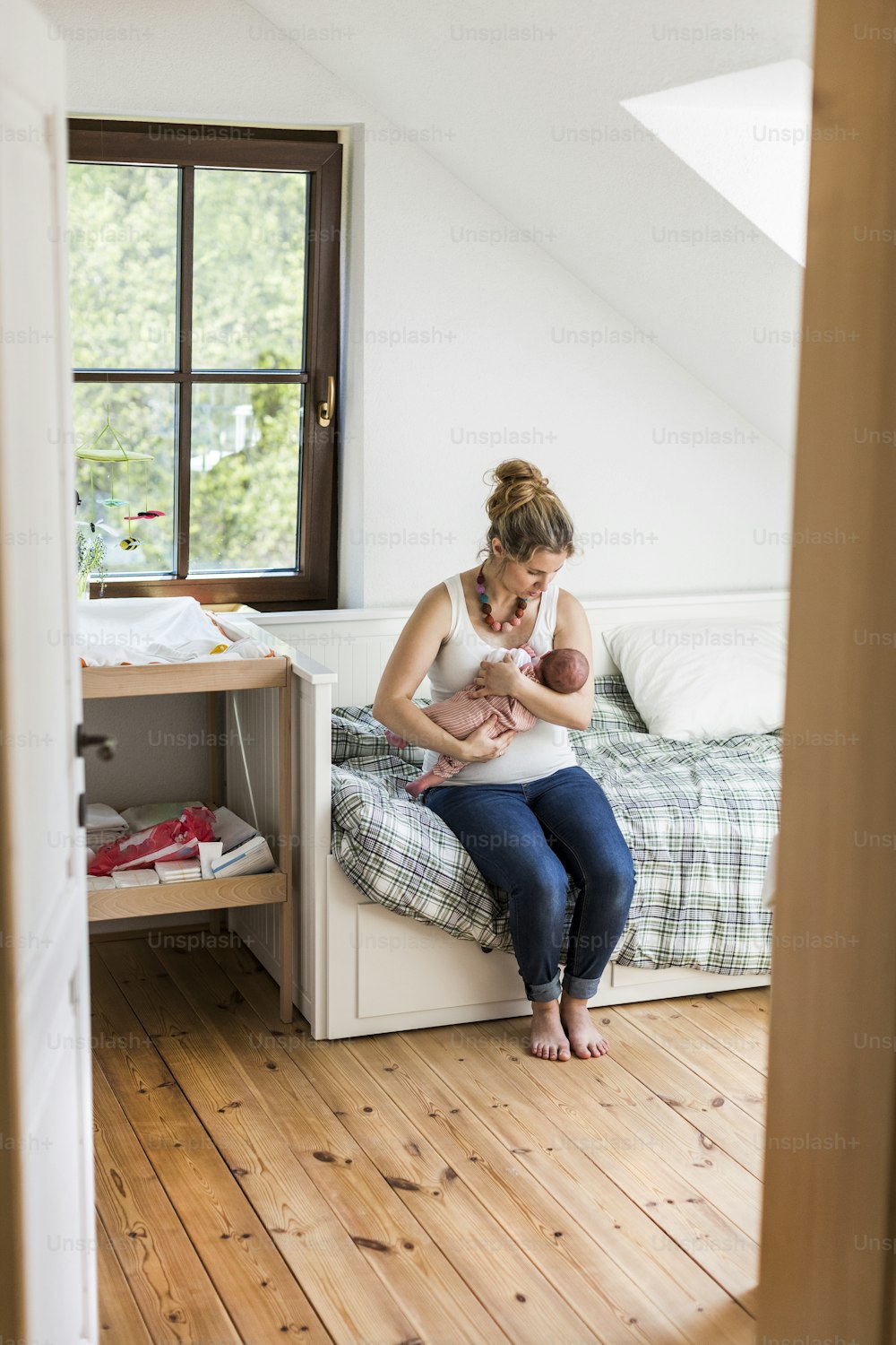 Beautiful young mother at home sitting on bed holding her cute newborn baby girl, kissing her on head.