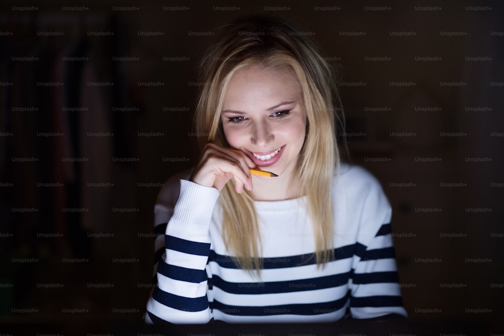 Young beautiful woman sitting at desk at night, wearing striped sweater holding pencil, smiling.