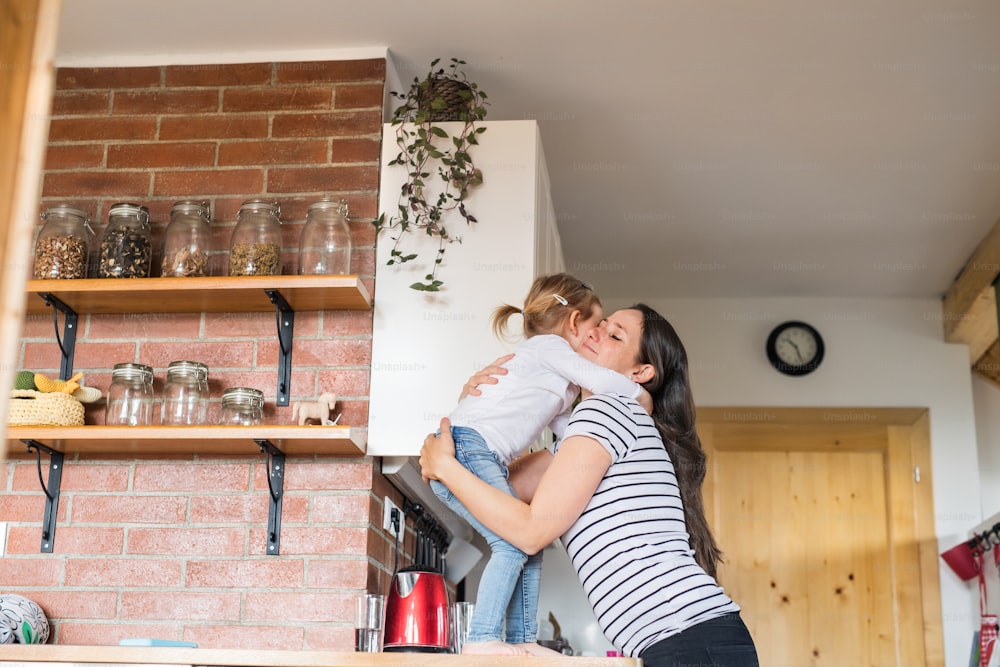 Linda jovem mãe em casa abraçando sua filhinha fofa em pé na bancada da cozinha.