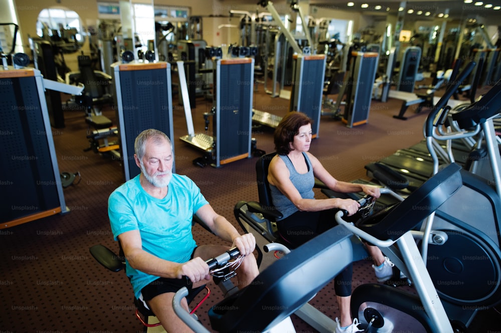 Beautiful fit senior couple in sports clothing in gym doing cardio workout, exercising on recumbent bicycle. Sport fitness and healthy lifestyle concept.