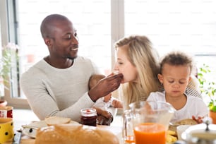 Hermosa familia joven interracial en casa con su linda hija y su pequeño bebé desayunando juntos.