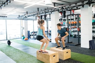 Beautiful young fit couple exercising in modern  gym, doing box jumps.