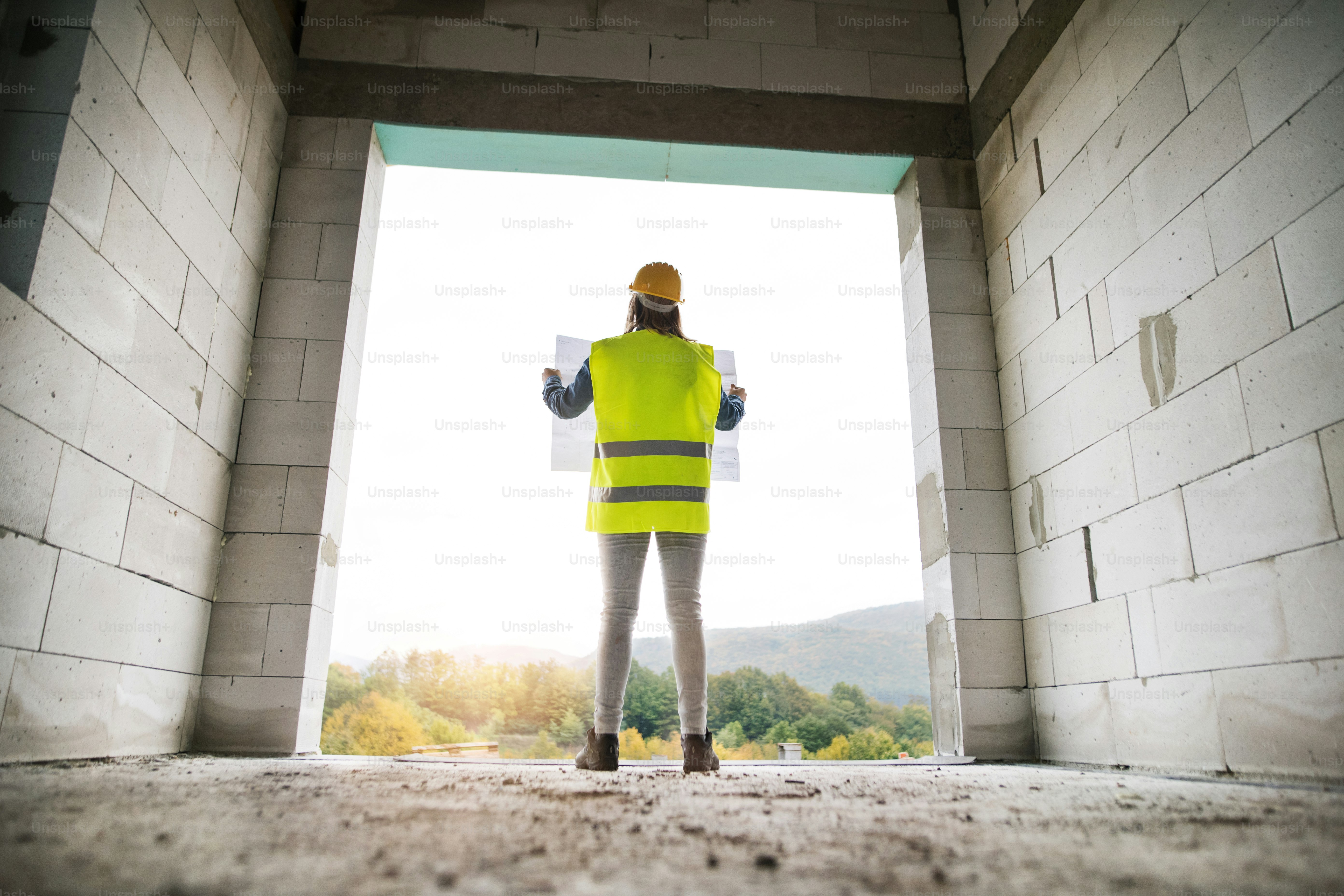 Arbeiterin auf der Baustelle. Schöne junge Frau mit Blaupausen. Hausbau. Rückansicht.