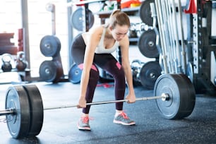 Beautiful young fit woman in gym lifting heavy barbell, flexing muscles