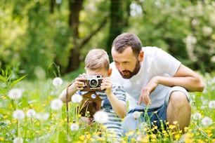 Young hipster father outdoors with his little son with camera, taking pictures of flowers. Sunny summer day.