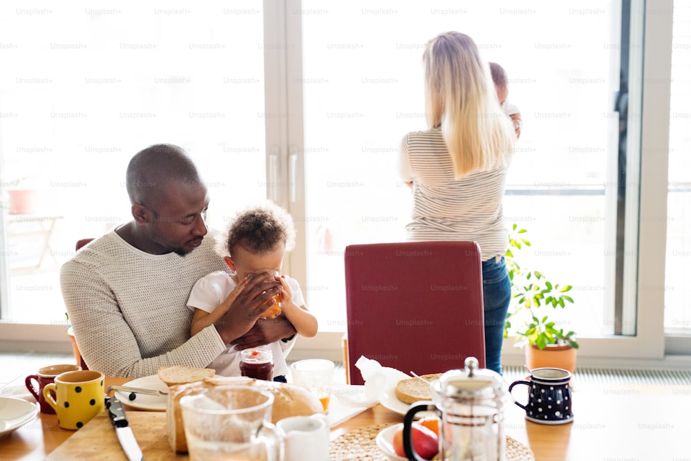 Linda família inter-racial jovem em casa com sua filha fofa e filho pequeno tomando café da manhã juntos. Mãe segurando menino nos braços, menina sentada no colo do pai, bebendo suco.