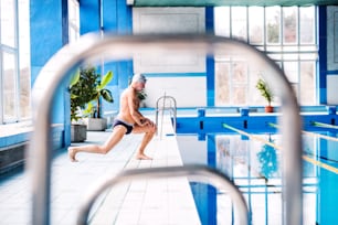 Senior man standing by the indoor swimming pool, stretching. Active pensioner enjoying sport.
