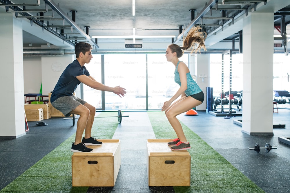 Beautiful young fit couple exercising in modern gym gym, doing box jumps.