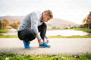 Handsome young hipster runner in gray jacket outside in colorful autumn nature, tying shoelaces.