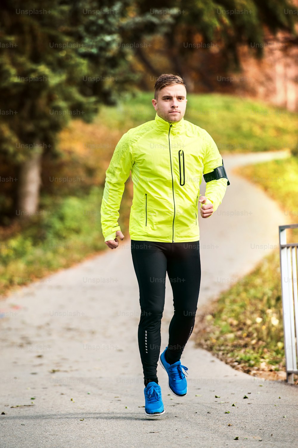 Young athlete with smartphone in yellow jacket running outside.Trail runner training for cross country running in colorful sunny autumn nature.