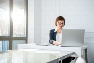 Élégante femme d’affaires senior vêtue du costume travaillant avec un ordinateur portable au bureau blanc