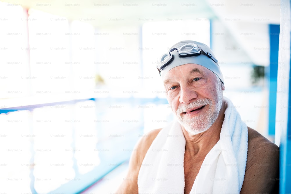 Senior man standing by an indoor swimming pool, a towel around his neck. Active pensioner enjoying sport.
