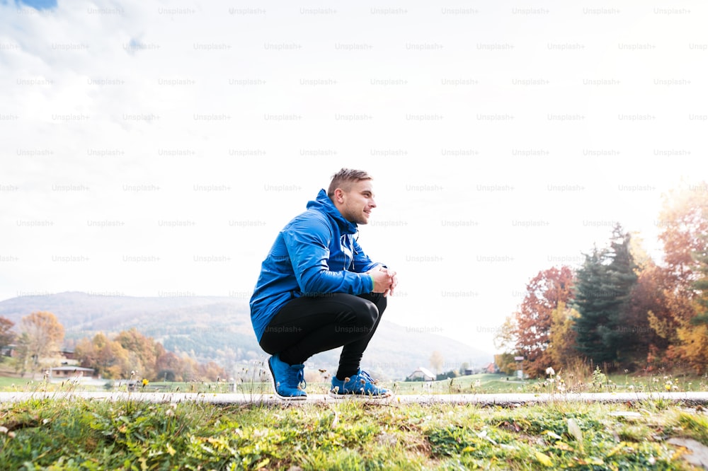 Young runner in blue jacket outside in colorful sunny autumn nature resting on an asphalt path. Trail runner training for cross country running.