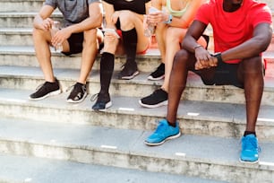 Jóvenes corredores irreconocibles en la ciudad juntos sentados en las escaleras, mirando un reloj inteligente o un teléfono inteligente, siguiendo su progreso en la carrera.