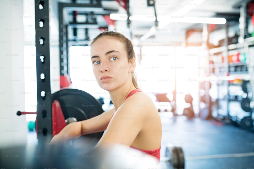 Beautiful young fit woman in gym standing at the heavy barbell, resting.